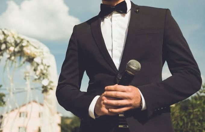 A man dressed formally, holding a microphone outdoors.
