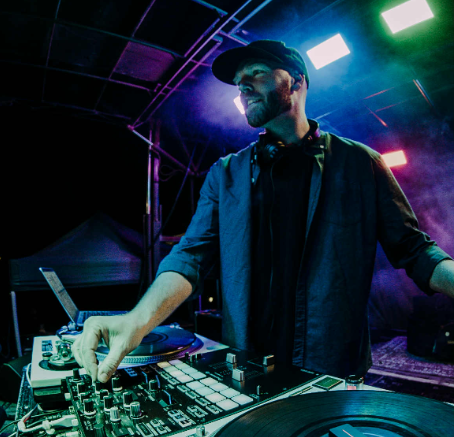 A man with a beard and a baseball cap is positioned behind the decks.