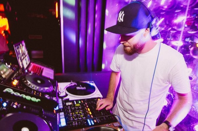 Avid DJ performing in a nightclub or music venue, bathed in dramatic purple light.
