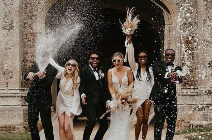 A wedding party posing in front of an old, arched stone building, possibly a church or manor house.